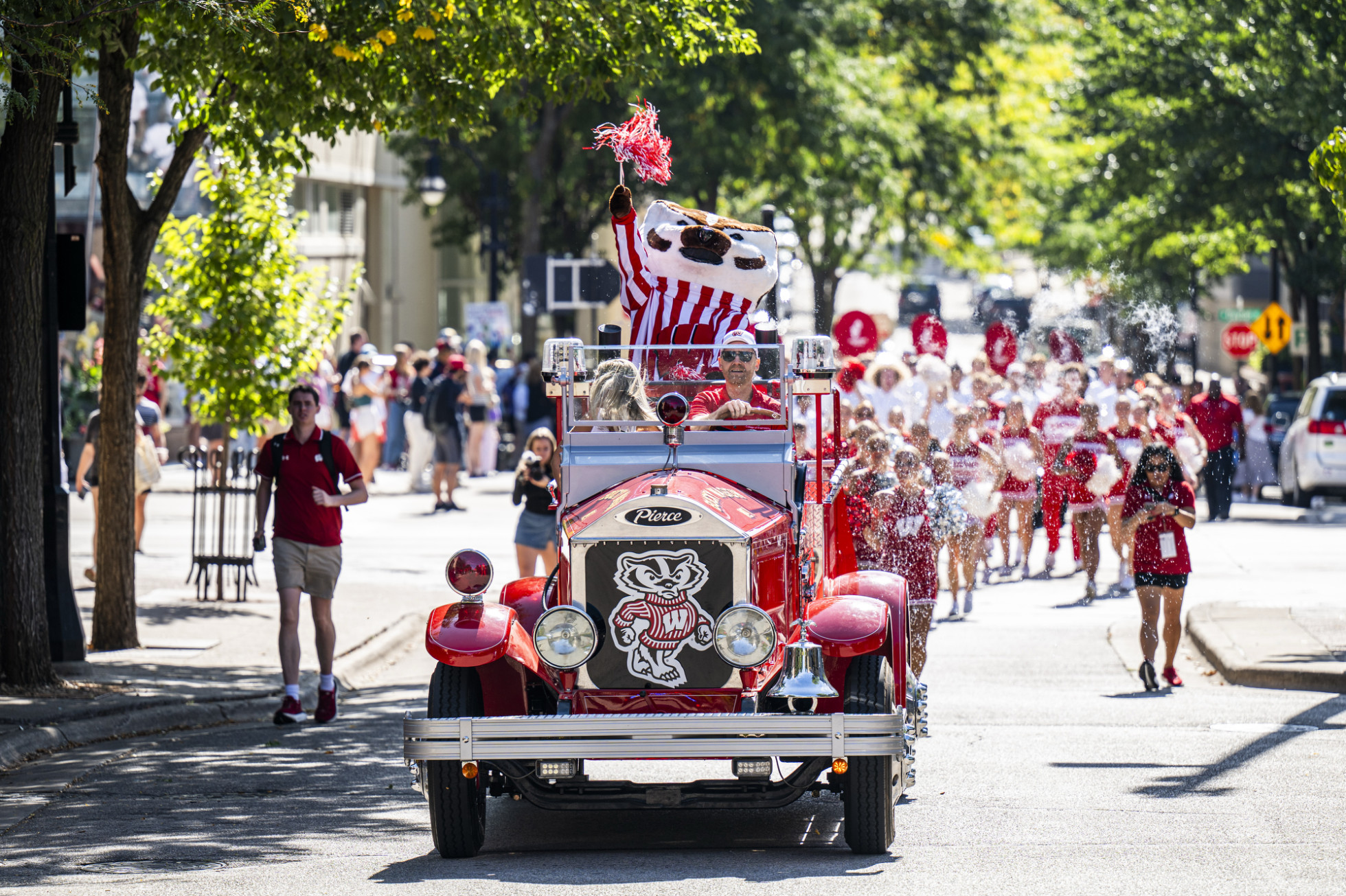 Bucky Badger, riding in the Bucky Wagon, waves a pom-pom in the air on State Street, followed by the UW Marching Band.