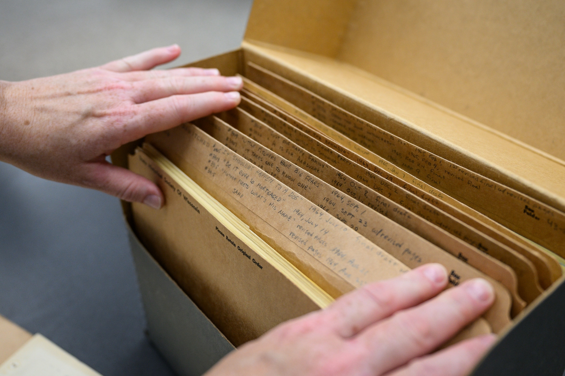 Hands looking through a box filled with brown file folders labeled as episodes of Bewitched from 1964-65.