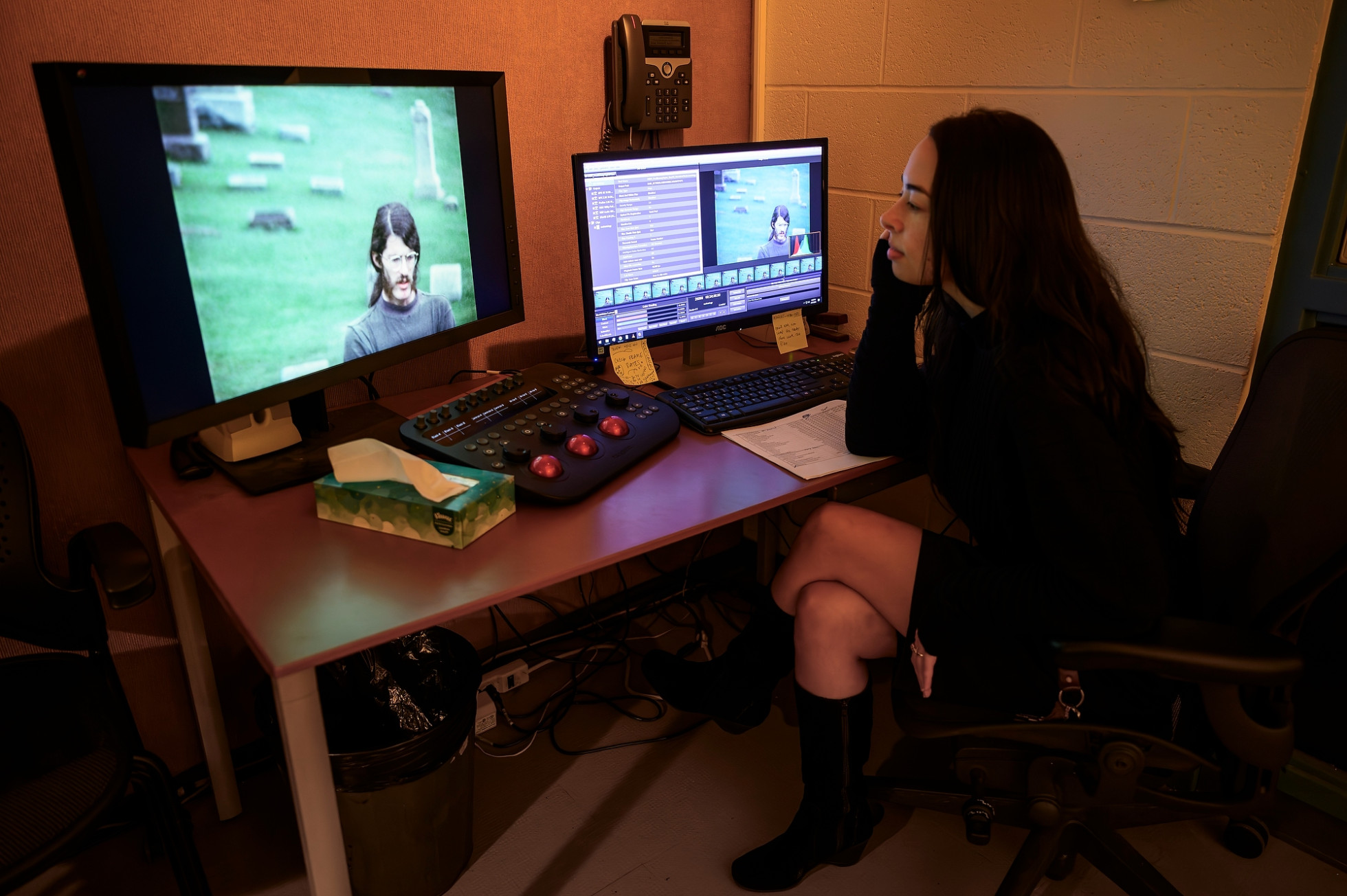A female student sits at a desk working with two computer monitors, one showing an outdoor video and the other displaying video editing software.