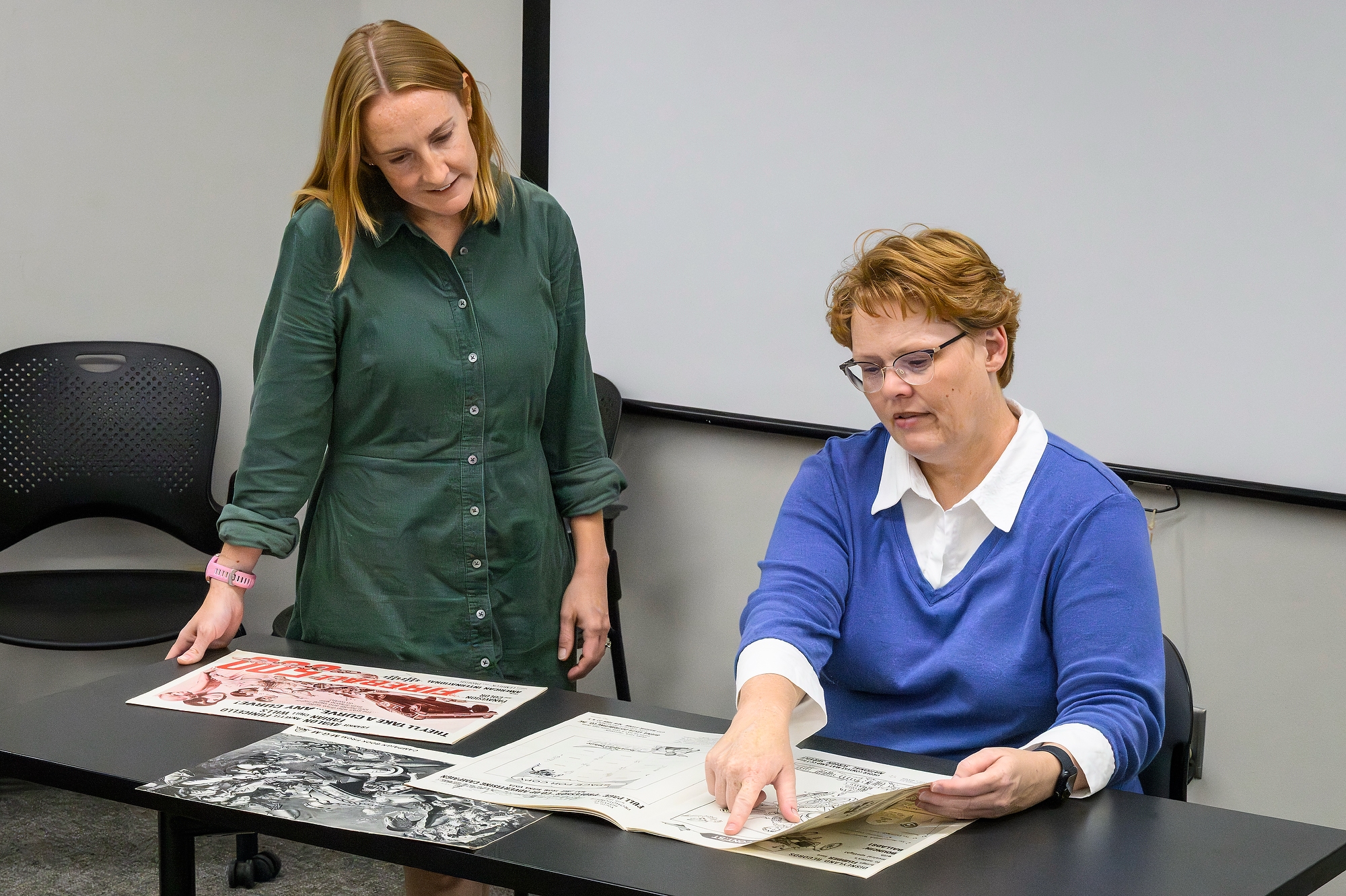 Amanda Smith, standing, and Mary Huelsbeck, seated, examine archival documents spread out on a table.