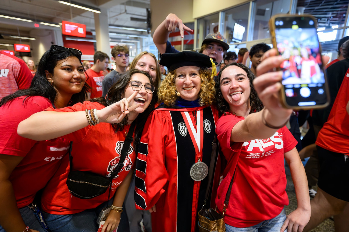 Three students pose with Chancellor Mnookin. One holds up her phone as they pose for a selfie.