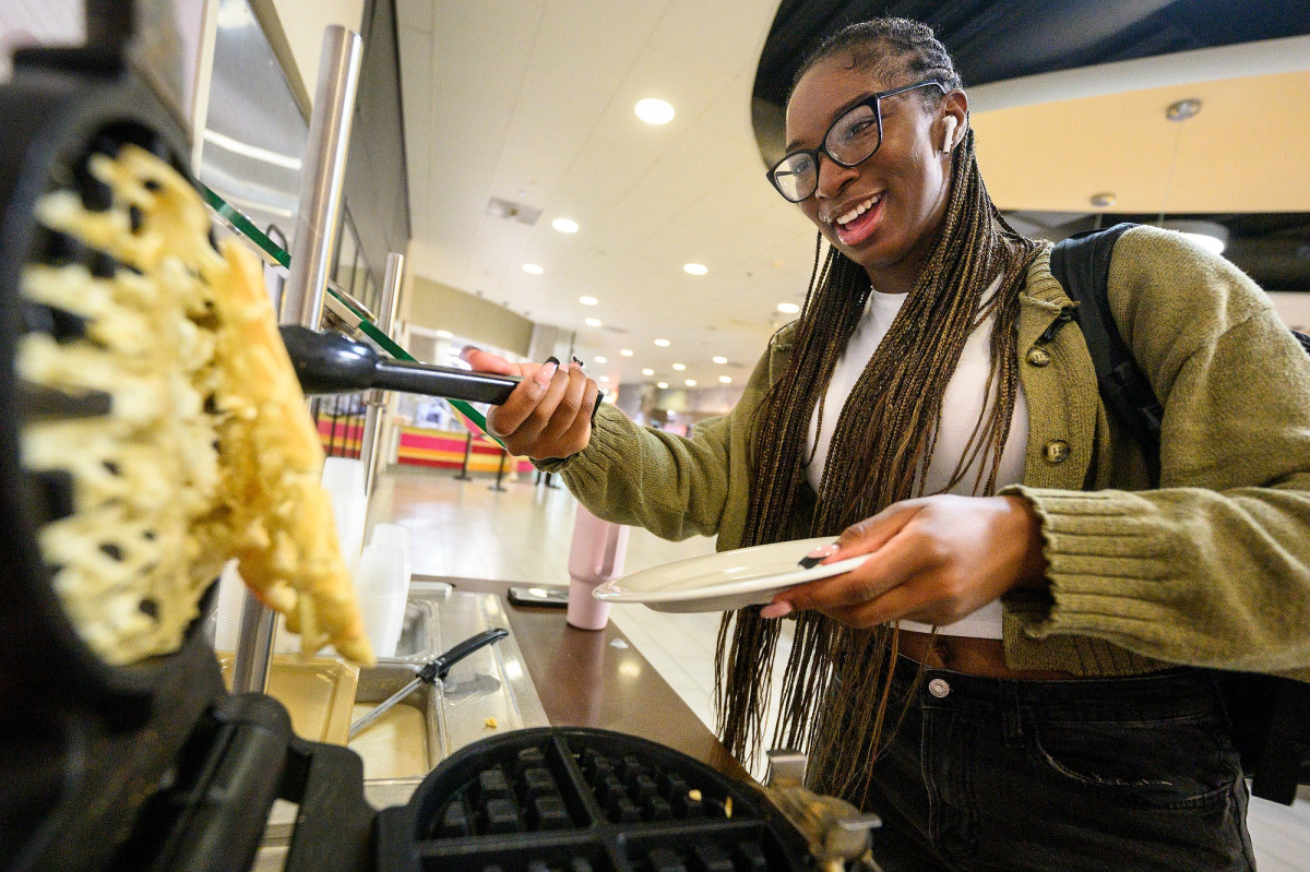 A student uses a waffle maker to prepare her breakfast. She smiles as she uses tongs to lift the waffle from the griddle.