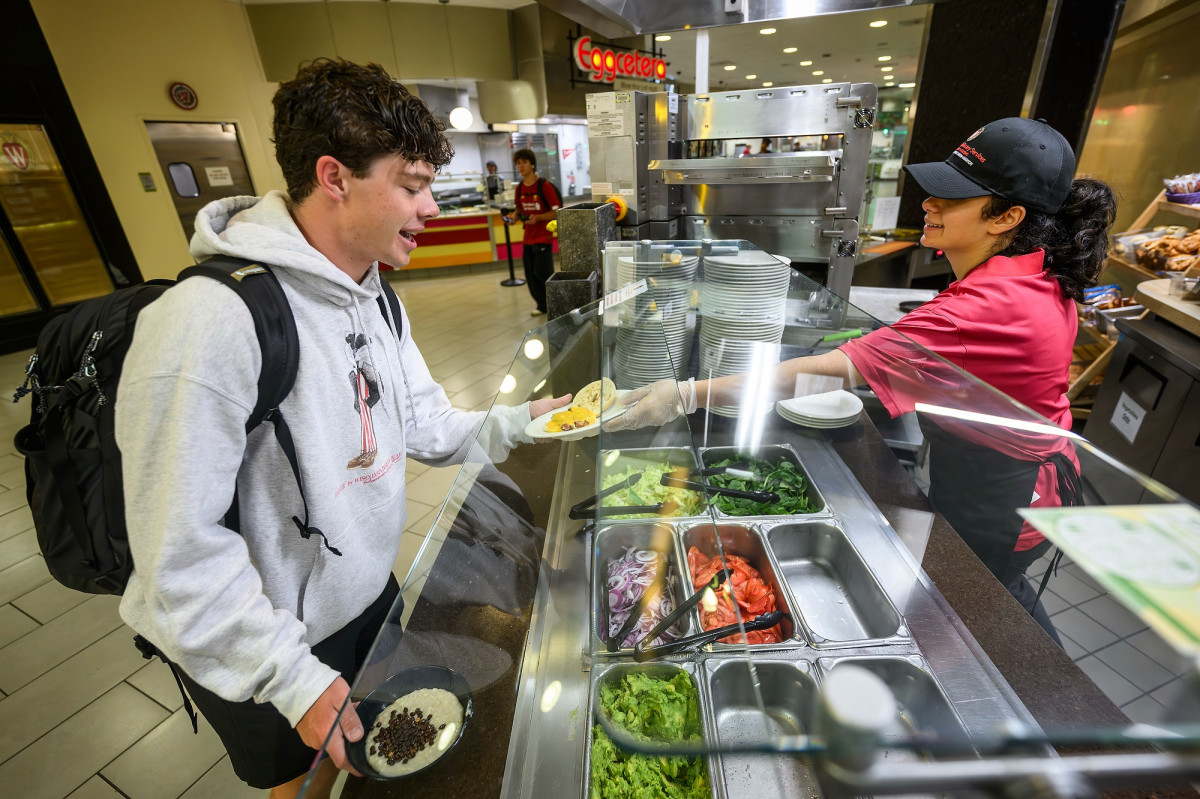 A student stands at a cafeteria buffet and receives a plate with an egg and bagel sandwich from a fellow student working the line.