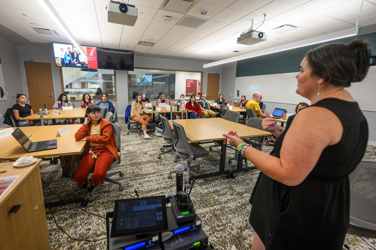 An instructor stands at the front of a classroom grouped around small tables and gives an introduction to the course.