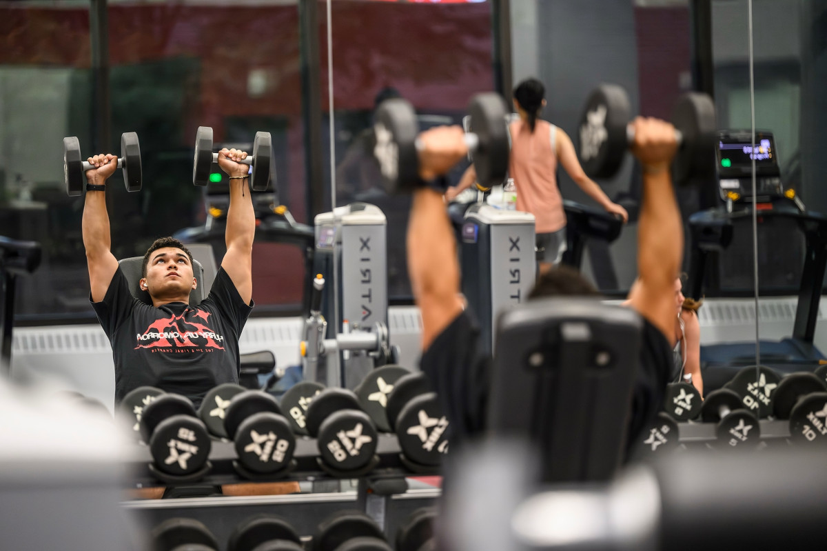 A student lies on a bench at the gym and lifts free weights over his head.