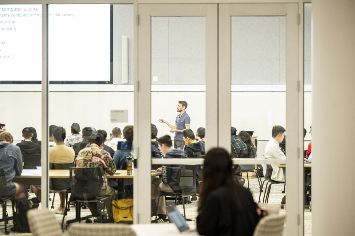 Seen through a glass door and wall, an instructor lectures to students seated at tables.