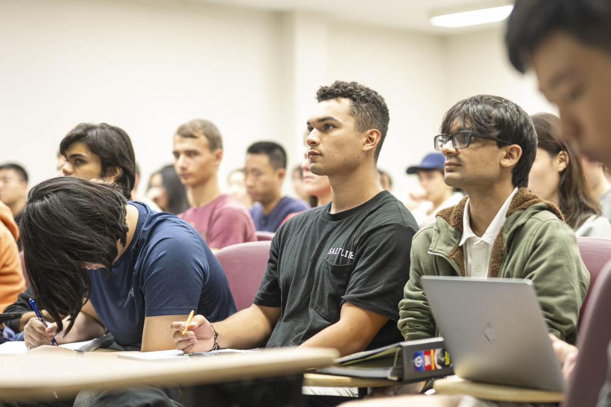 Borrero sits surrounded by students who sit in a lecture hall and listen to someone speaking at the head of the room. 