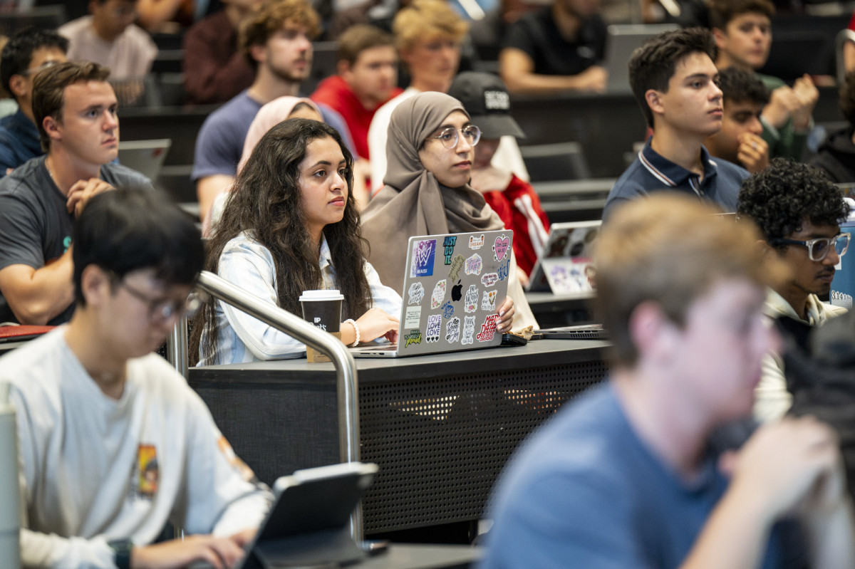 Students sit in a lecture hall and listen to someone speaking at the head of the room. 
