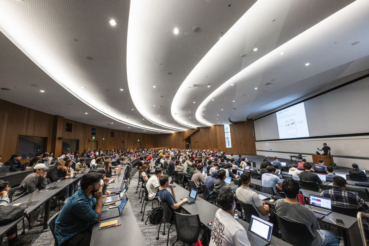 Students sit in a lecture hall and listen to someone speaking at the head of the room. 