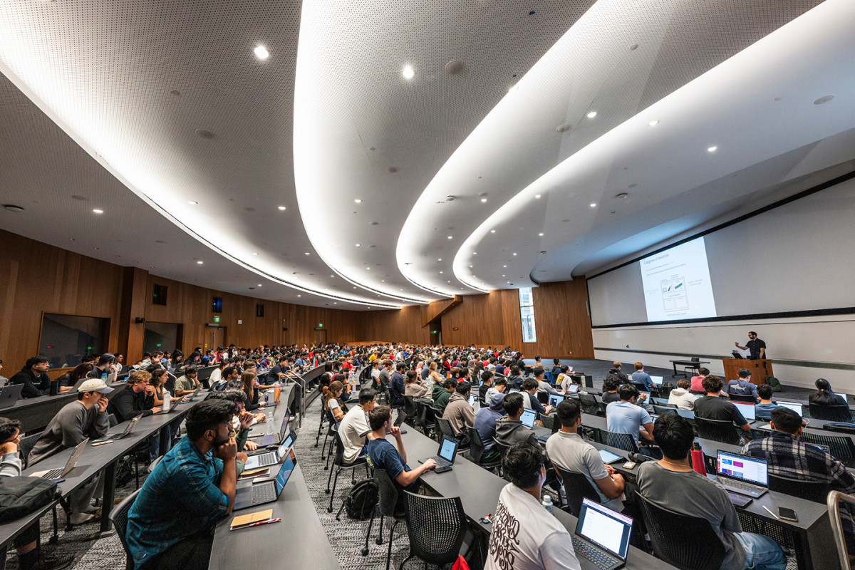 Modern classroom with many students sitting in rows with laptops facing and instructor who is lecturing behind a podium