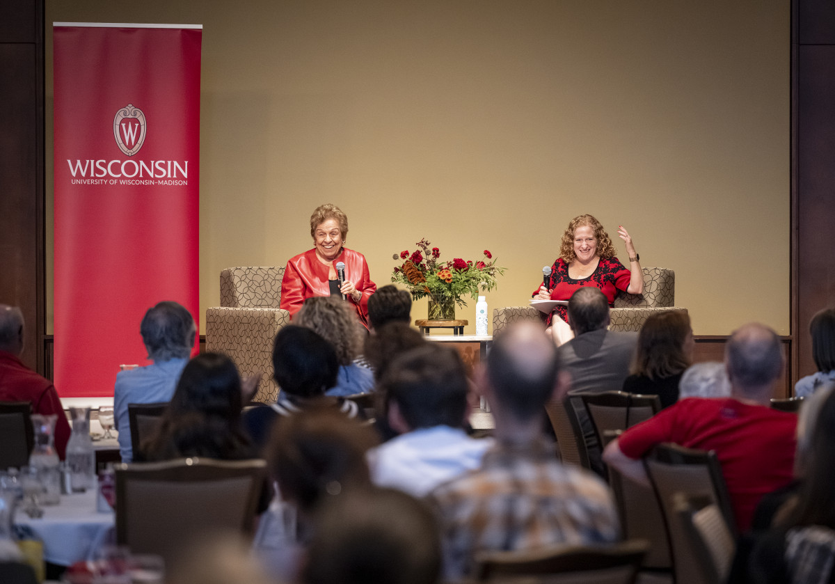 Shalala and Mnookin sit in armchairs on a stage before an audience seated at banquet tables. They're smiling and gesturing as they speak.
