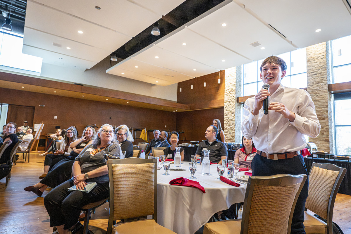 A student stands among the audience and smiles as he asks a question.