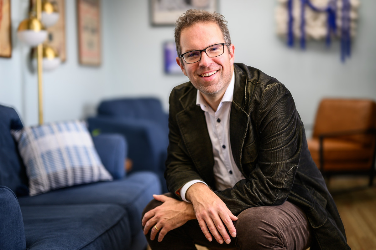 Eric Hoyt, wearing glasses and a dark jacket, leans forward and smiles in a comfortable-looking room with a couch and chairs, and framed archival artwork on the walls.