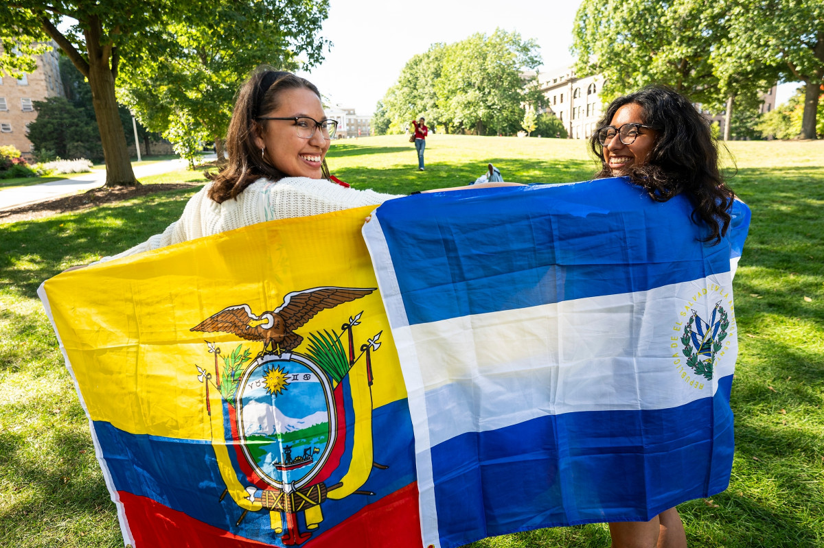 Two women drape flags representing their Latine heritage actoss their backs. On the left, a woman holds the flag of Ecuador. On the right, a woman holds the flag of El Salvador. They're standing in the grass at the bottom of Bascom hill and smiling to the camera.