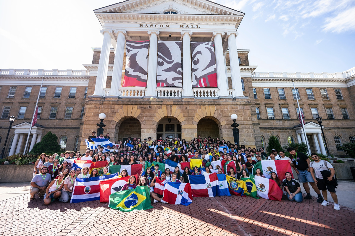 A group of nearly 300 gather outside the entrance to Bascom Hall. They're holding up national flags representing their Latine heritage as they stand under a large banner of Bucky Badger.