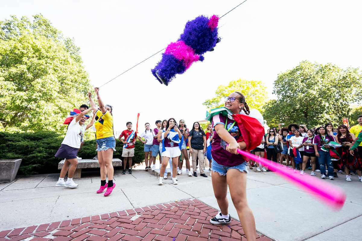 A student takes a big swing at a piñata while a gathered crowd stands and cheers.