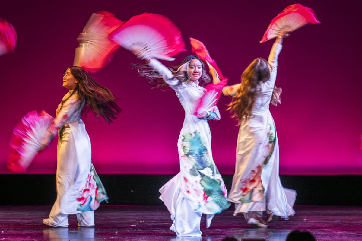 Three student wearing Vietnamese cultural attire dance to a song while holding fans in their hands. 