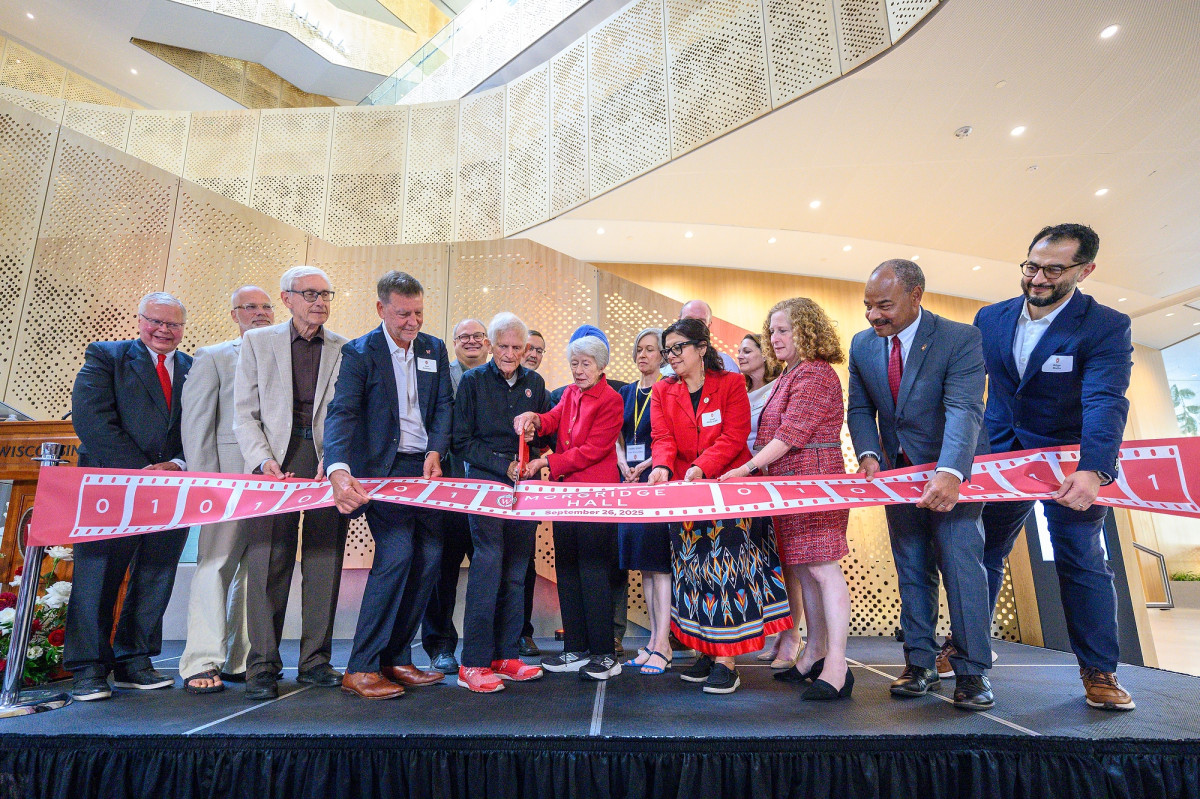 A group of people stand on stage. The Morgridges hold a large pair of red scissors in their hands as they cut a red ribbon that is displayed in front of the large group.