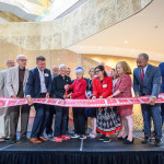 A group of people stand on stage. The Morgridges hold a large pair of red scissors in their hands as they cut a red ribbon that is displayed in front of the large group.