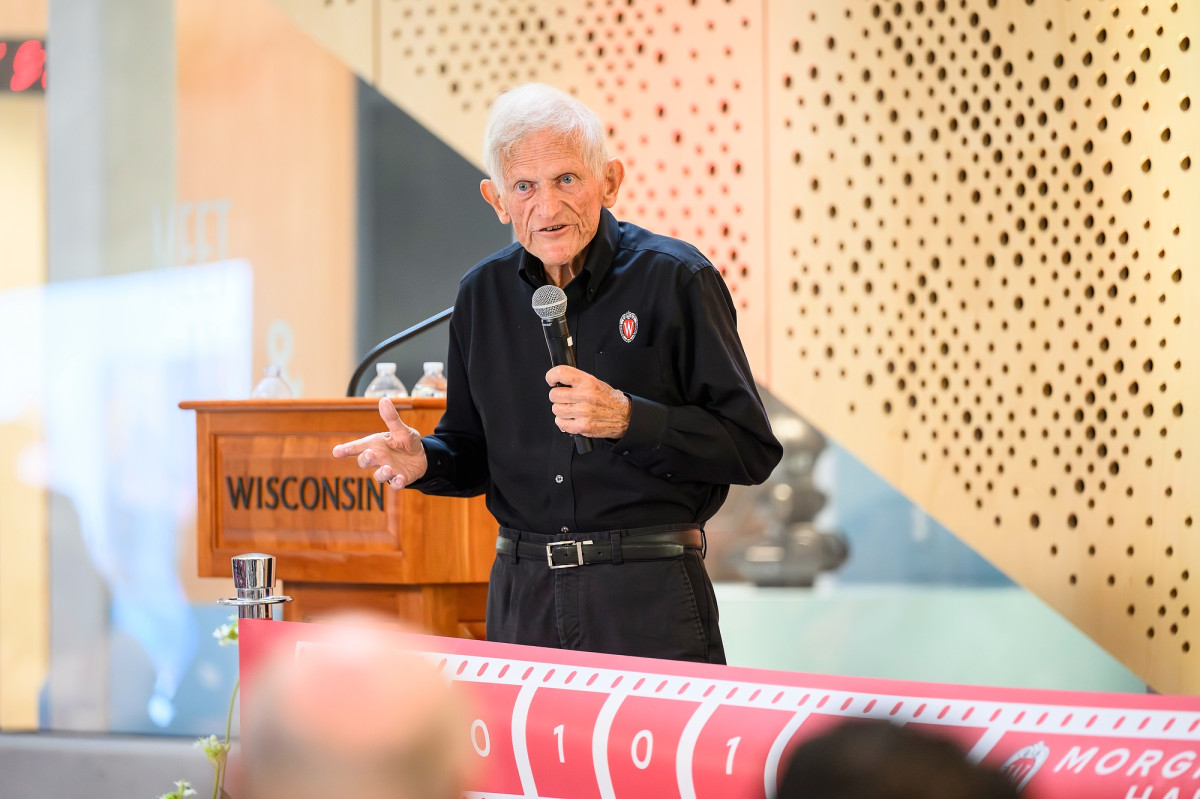 John Morgridge stands next to a podium and holds a microphone as he speaks to a crowd.