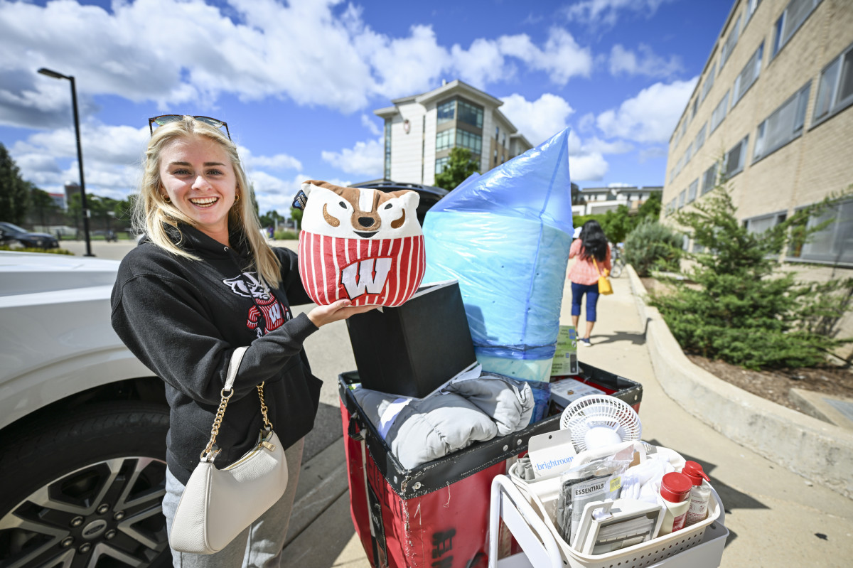 A students stands next to a red move-in card. She holds a stuffed animal in the shape of Bucky Badger and smiles at the camera. 