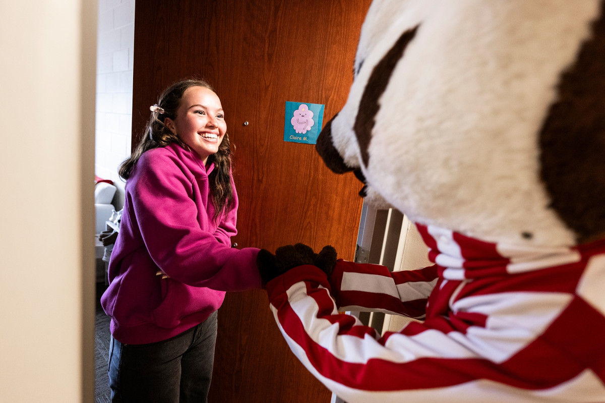 A students stands in the doorway of her residence hall room and shakes Bucky Badger's hand. 