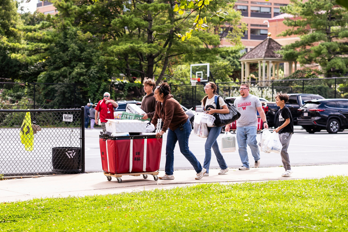 A person pushes a red move-in cart filled with belongings while being followed by several people who also carry goods. 