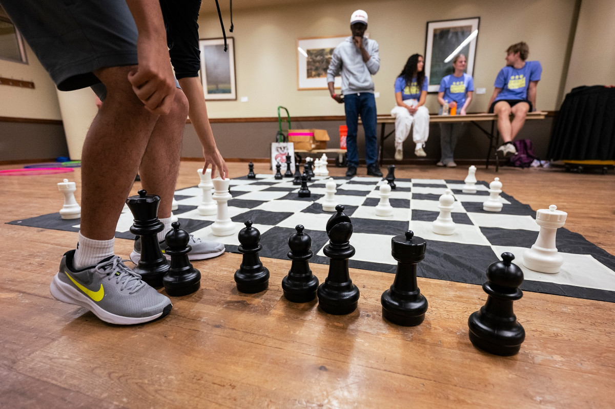 A giant chessboard fills the floor of the room. Two students stand on either side in the midst of the challenge. 