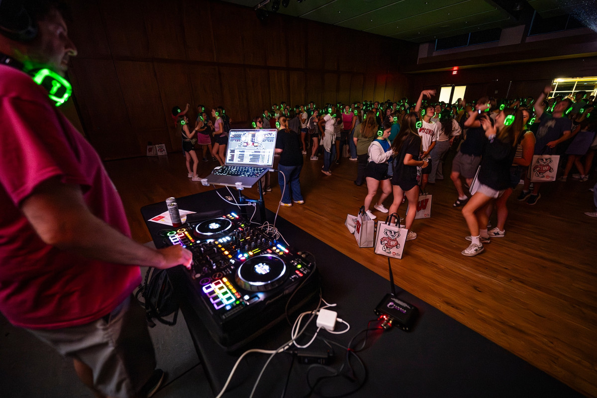A DJ plays to a crowded room filled with students listening to headphones at a silent dance party. 