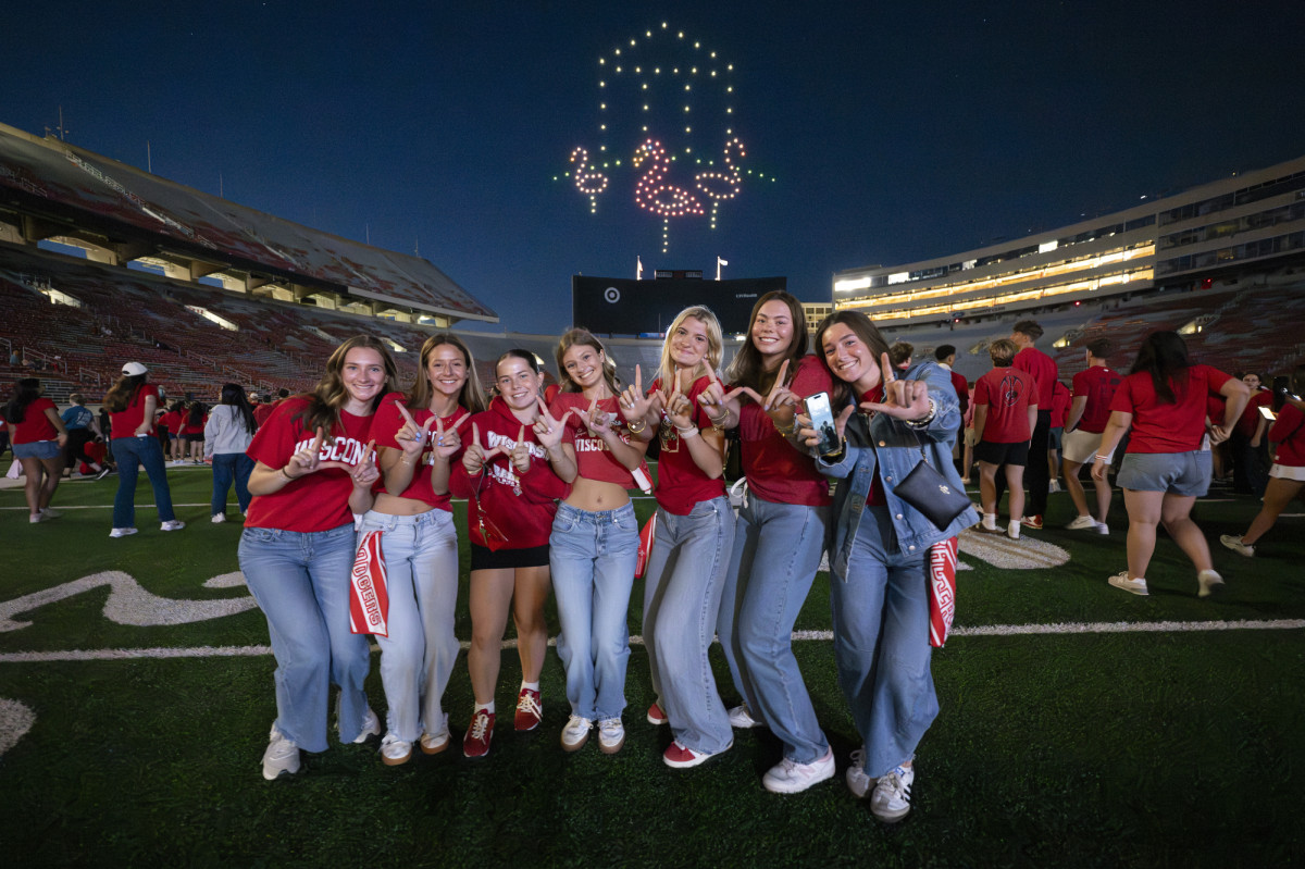 A group of students stand on the field at Camp Randall Stadium and flash a 'w' with their hands. 