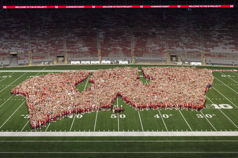Thousands of UW students stand on the football field at Camp Randall Stadium, most wearing red t-shirts, forming the shape of UW Athletic's Motion W logo.