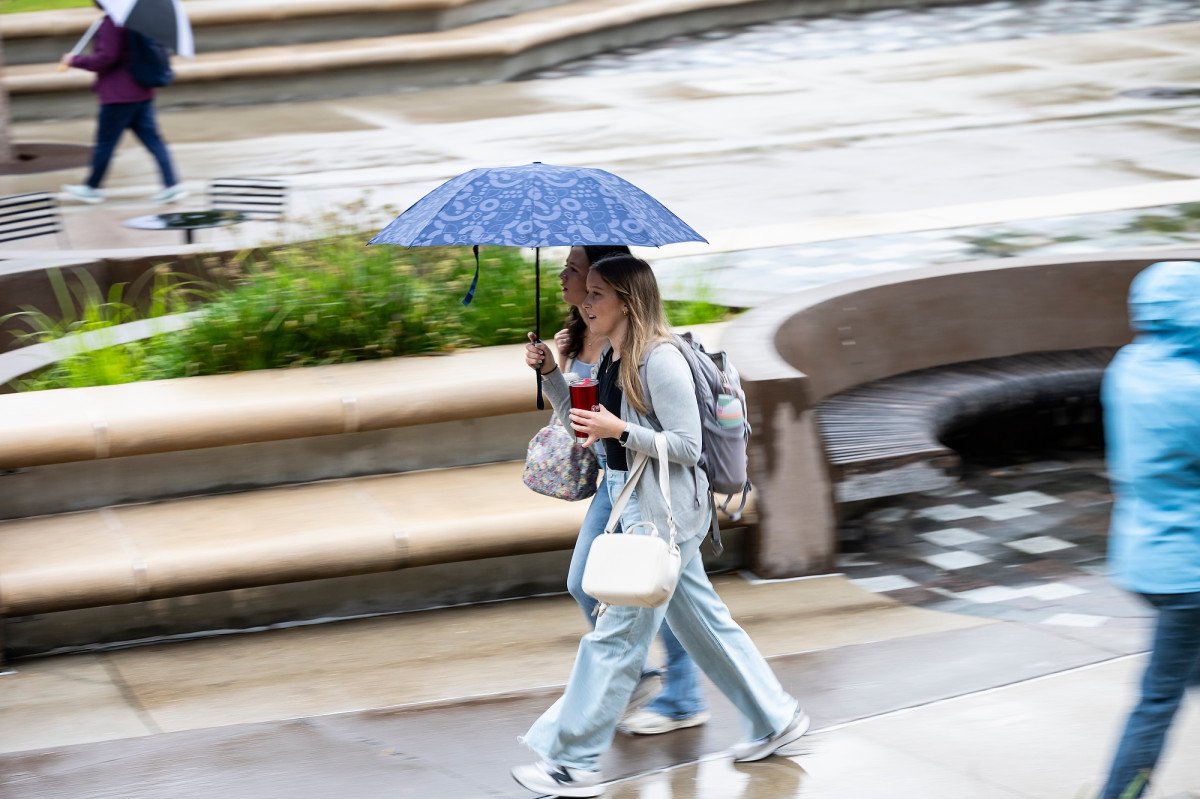 Two students huddle together under an umbrella as they walk together down a pedestrian mall.