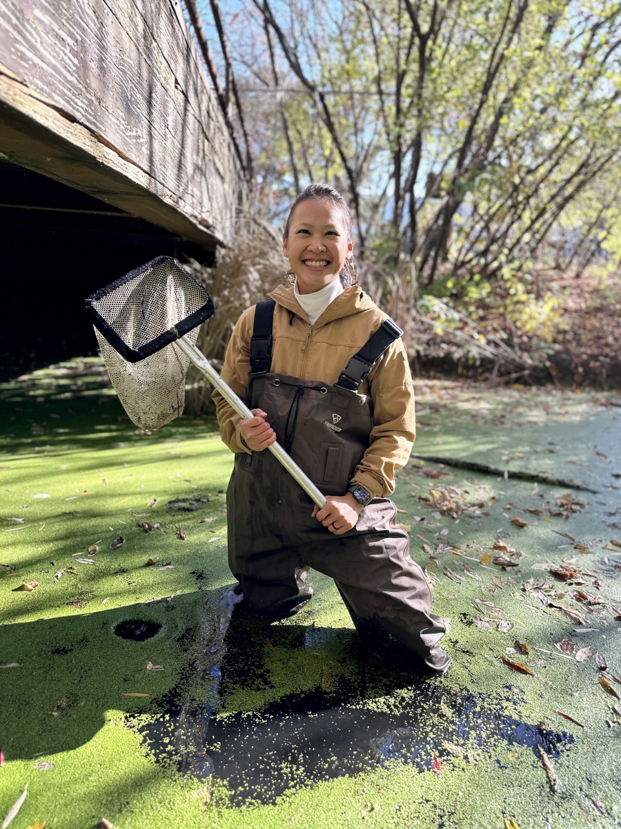 Jessica Hua wears waders as she stands smiling in knee-deep water holding a sampling net.