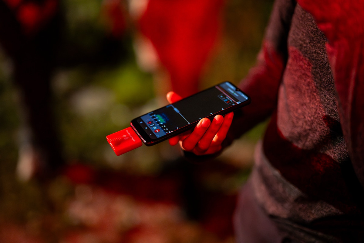 Close-up of a hand holding a phone with a small device attached. The screen shows an echolocation app open.