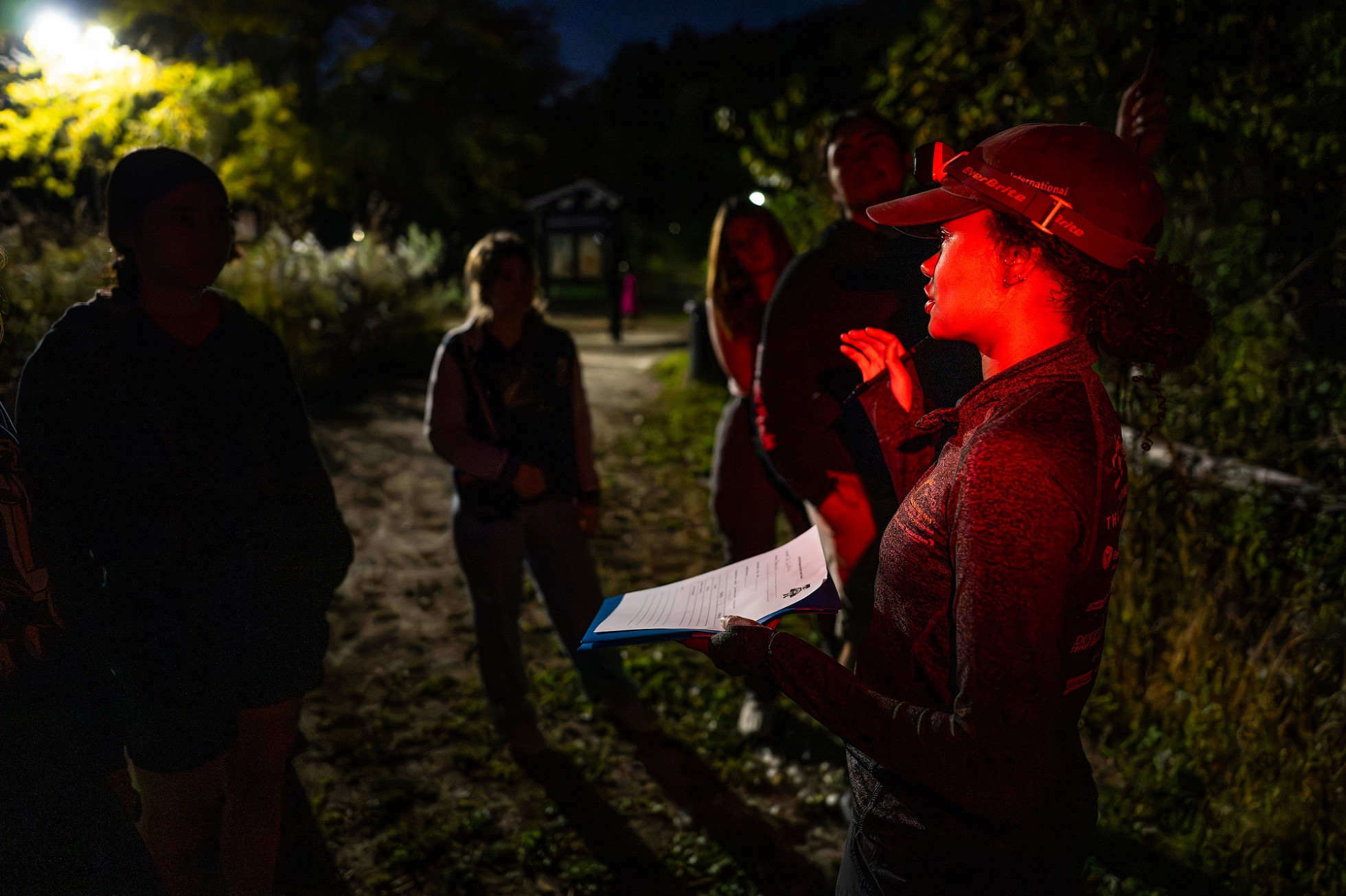 Students wear red light headlamps and gather on a wooded path to talk about bats