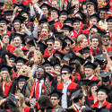 A joyful group of UW-Madison graduates in black caps and gowns with red accents celebrate together, smiling and raising their hands.