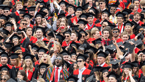 A joyful group of UW-Madison graduates in black caps and gowns with red accents celebrate together, smiling and raising their hands.