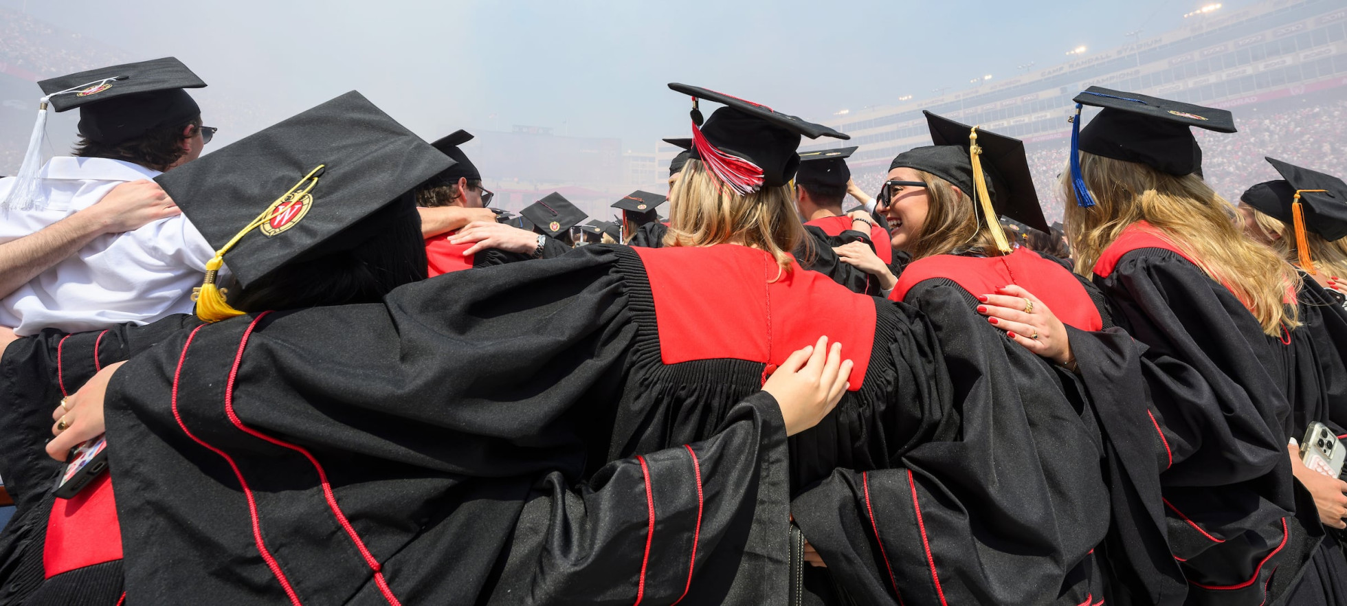 A group of UW graduates in black gowns with red accents stand arm-in-arm at Commencement in Camp Randall Stadium as they celebrate their achievement together.
