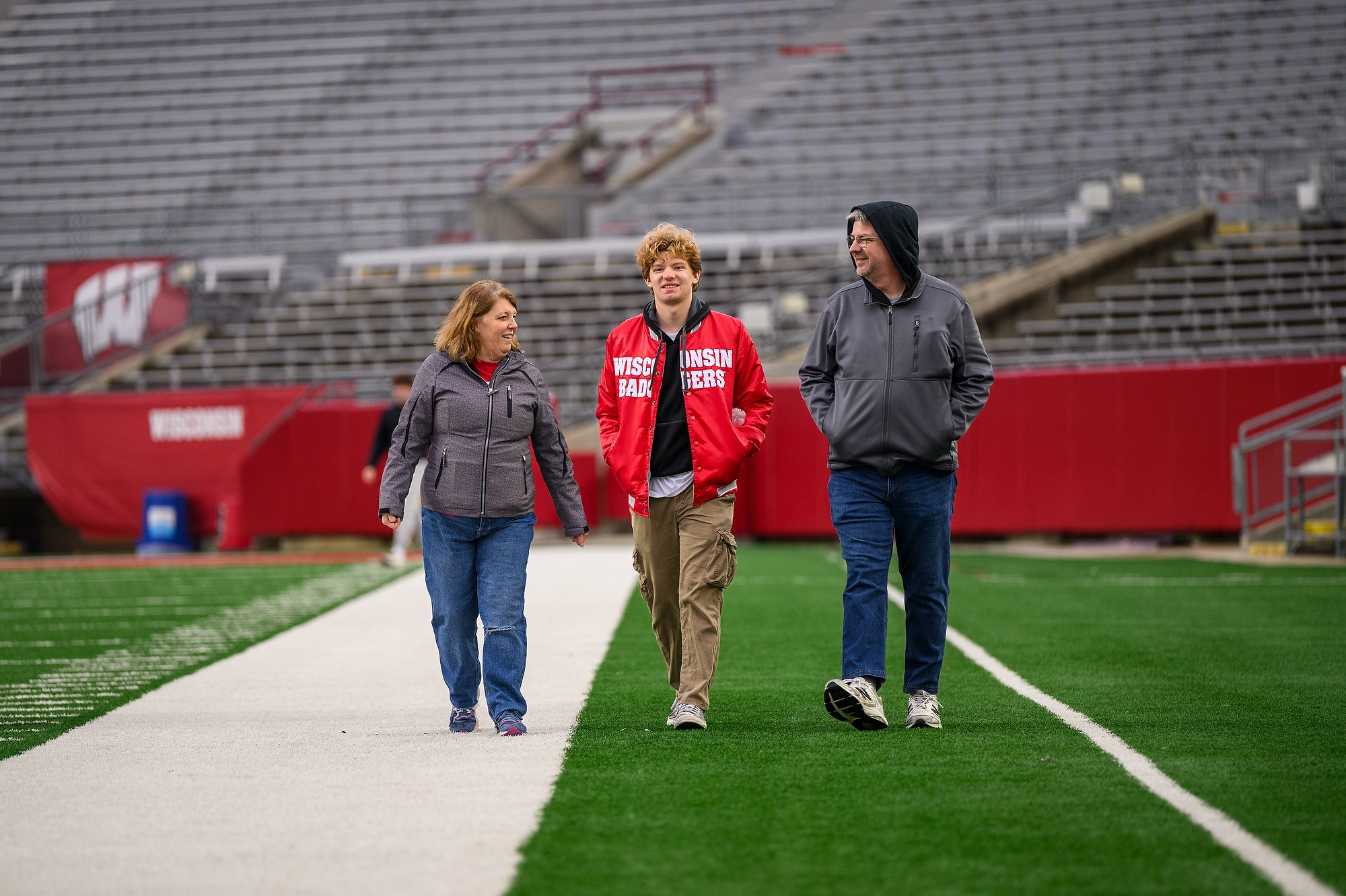 A student wearing a red Wisconsin Badgers jacket walks down a football field between his parents.