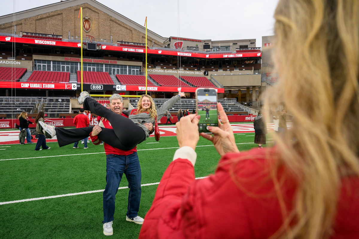 A woman takes a photo of a man holding his daughter, who is posing in his arms in front of the football field end zone.