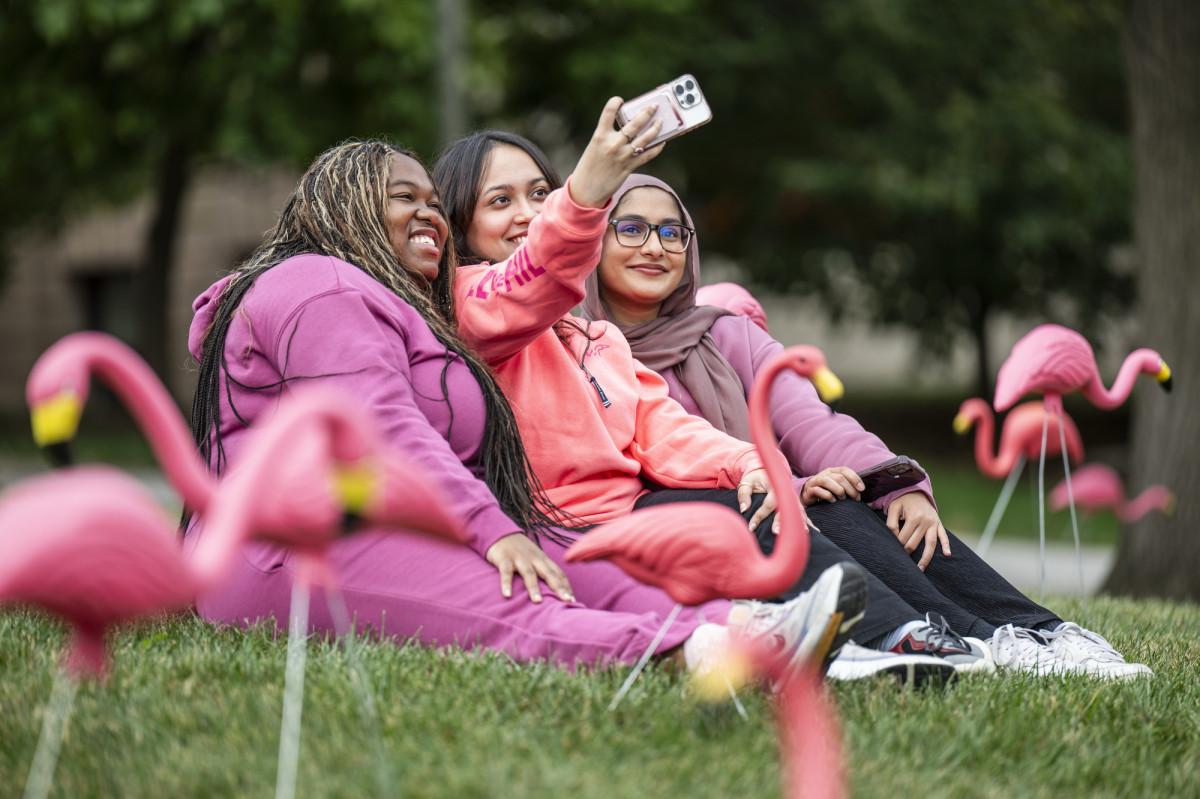 Three students wearing pink sit on among the flamingos on Bascom Hill. One of the girls holds up a phone to take a selfie of the small group.