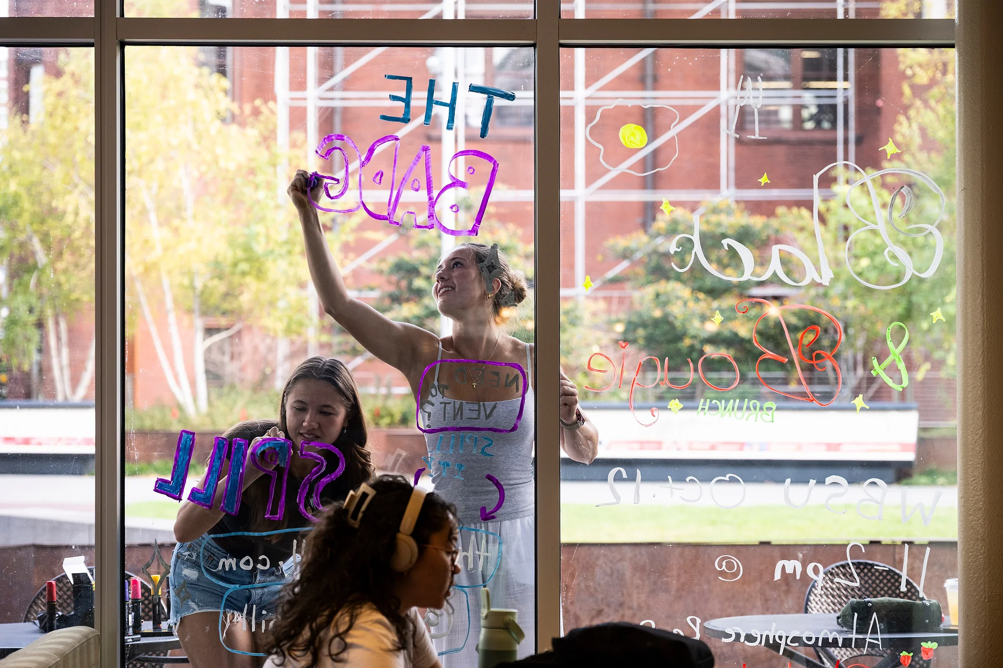 From inside Memorial Union, a group of students are shown drawing designs on the windows of the building's Lakeshore Lounge.