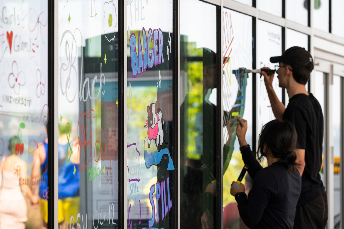 Two students are shown holding large window markers and draw designs on the windows of Memorial Union.