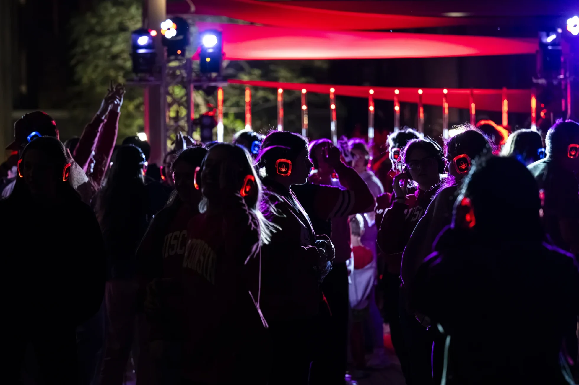 A crowd of people silhouetted against colored stage lights dance while wearing glowing headphones
