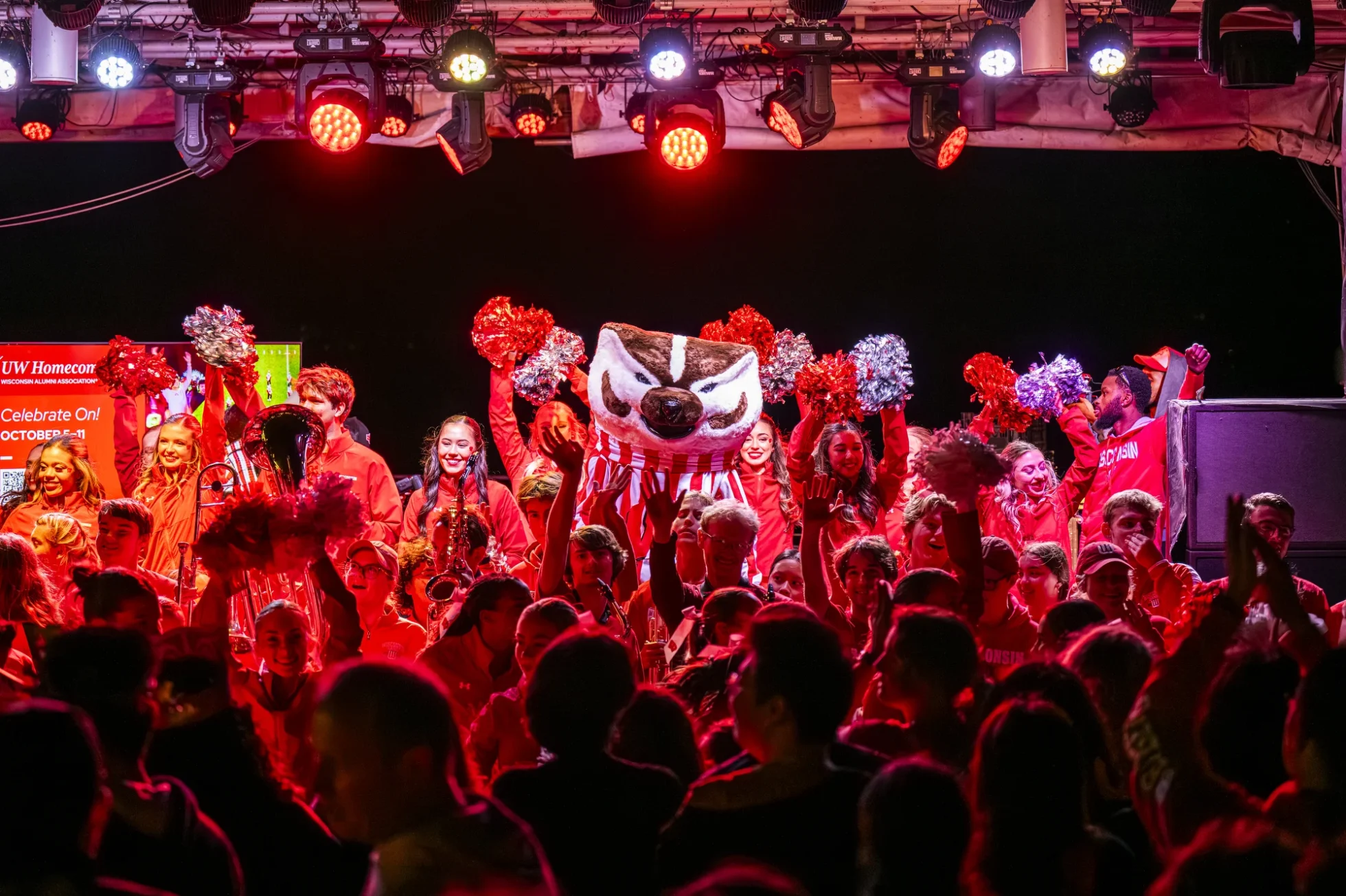 Bucky Badger and Spirit Squad members holding pom poms line a stage while people in a crowd cheer