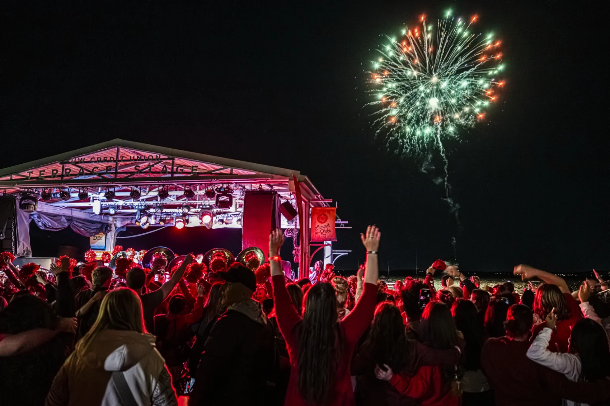 A large crowd of people cheer as fireworks explode over Lake Mendota at night and the UW Marching Band performs near the Memorial Union outdoor stage
