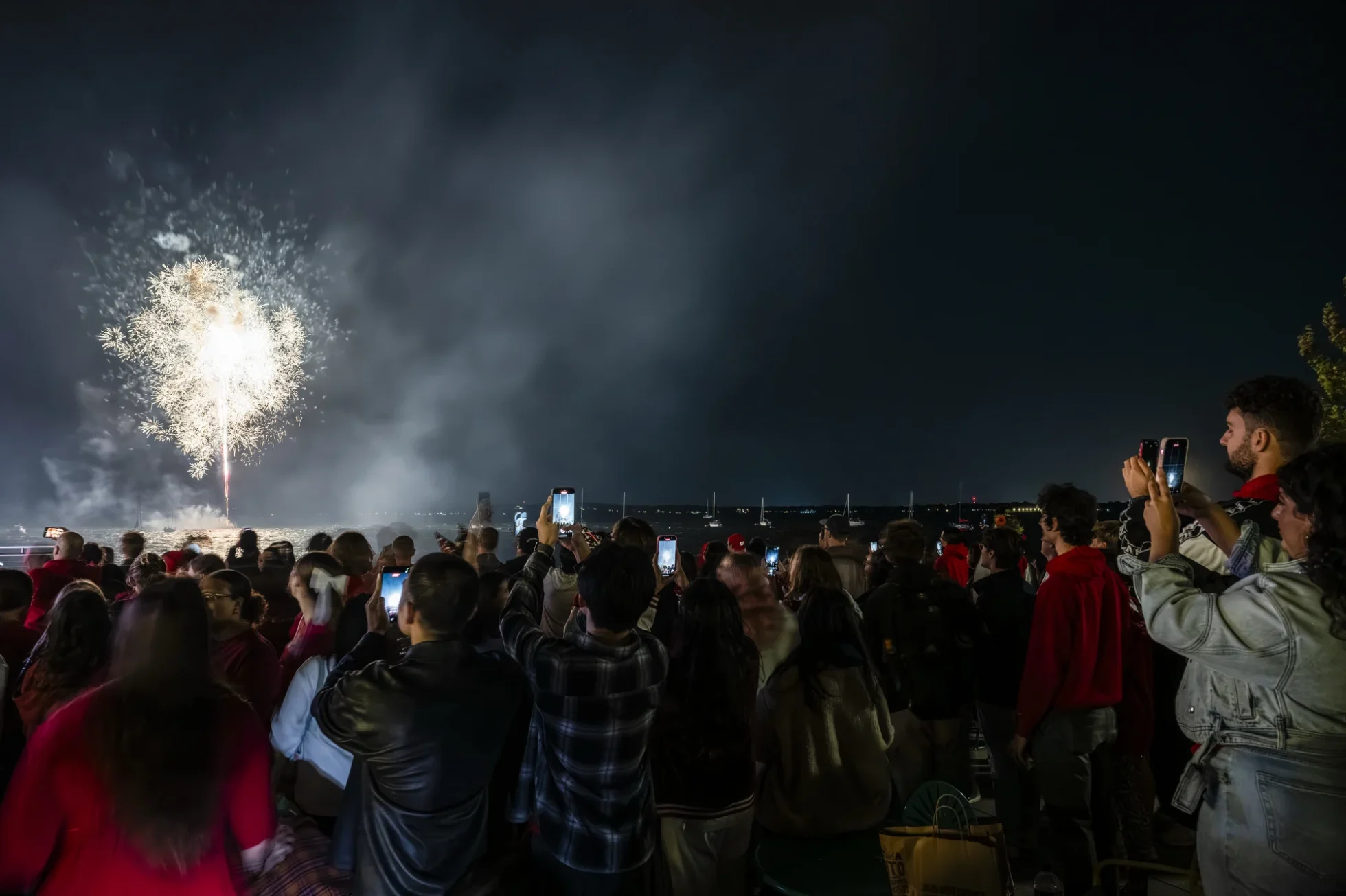 A large crowd of people watch and record with their phones as fireworks explode over Lake Mendota at night