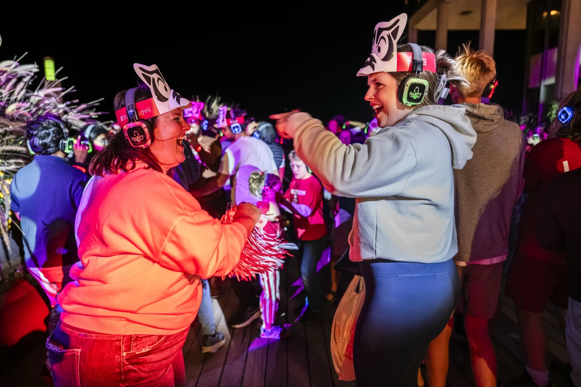 Two women wearing paper Bucky hats and illuminated headphones smile and dance together amid colorful lights