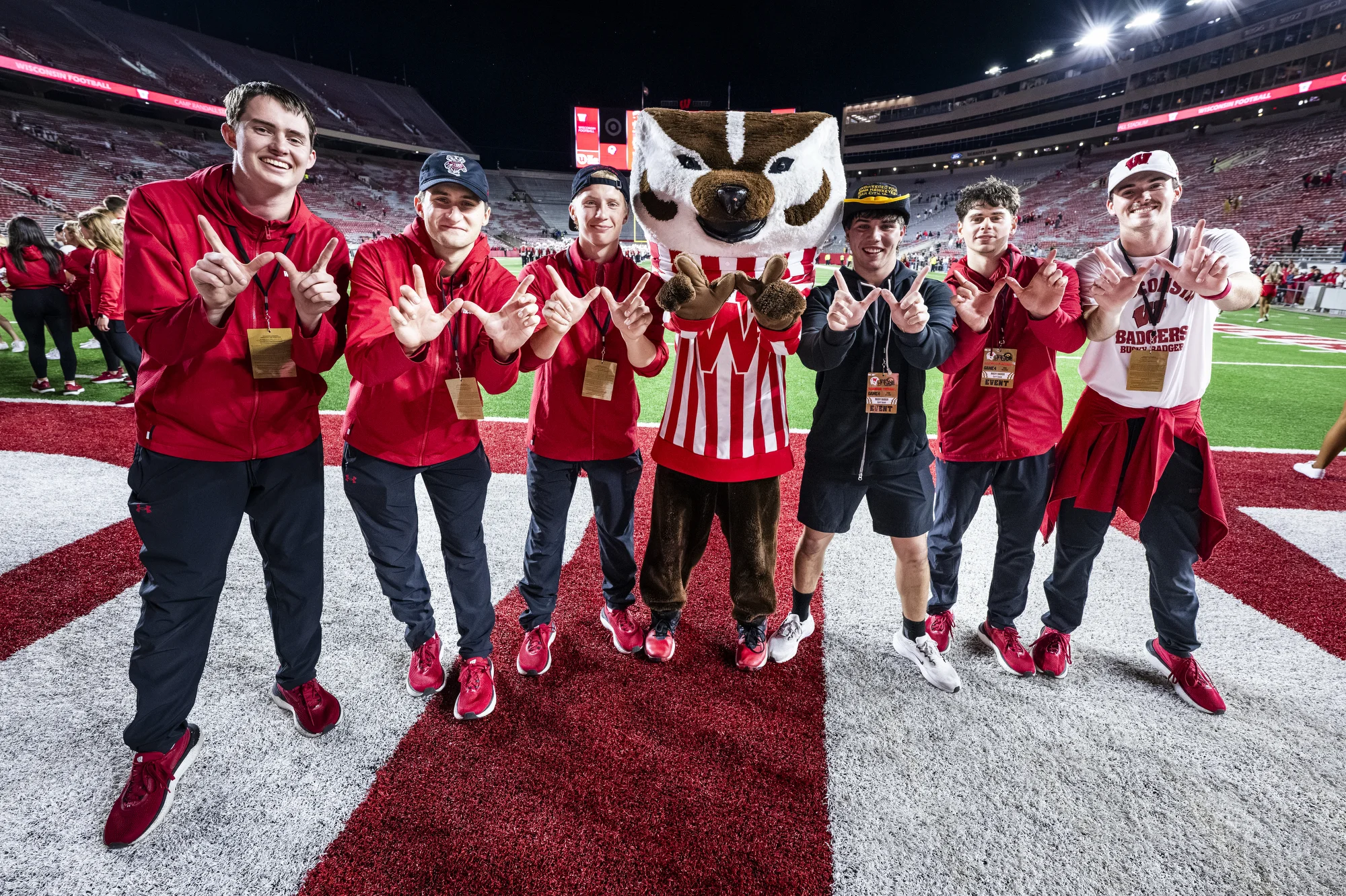 Current and former Bucky Badger performers throw "w" signs alongside the mascot on Camp Randall field