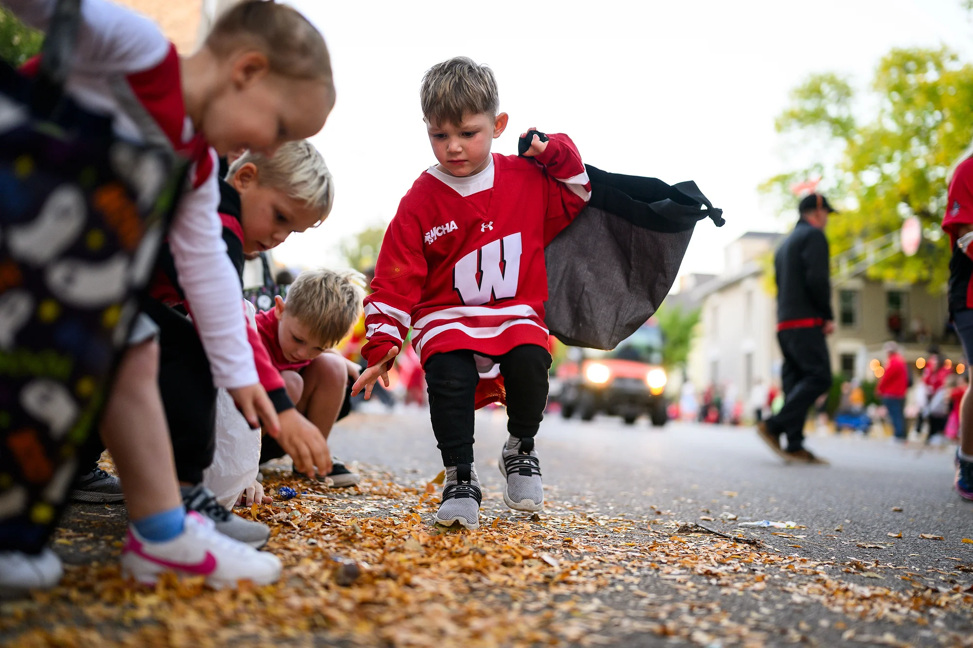 Children wearing Badger gear collect candy from a leaf-covered street during the Homecoming parade.